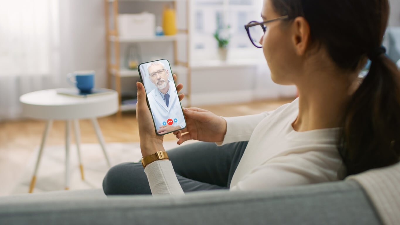 A young woman sits at home on her phone, talking to her doctor over video call.